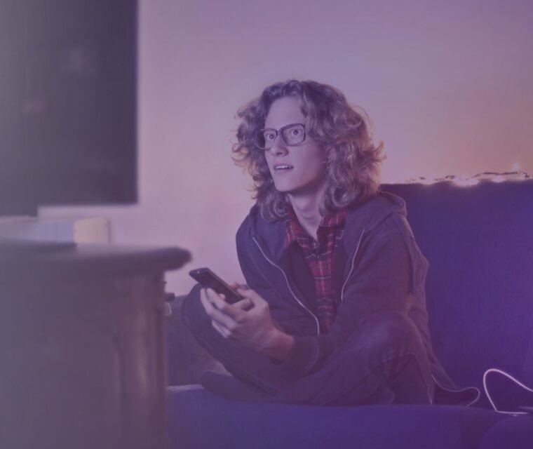 Young man in a living room using a smartphone and watching TV, enjoying seamless streaming with the best IPTV service and premium channels. - iptv portugal - united kingdom iptv